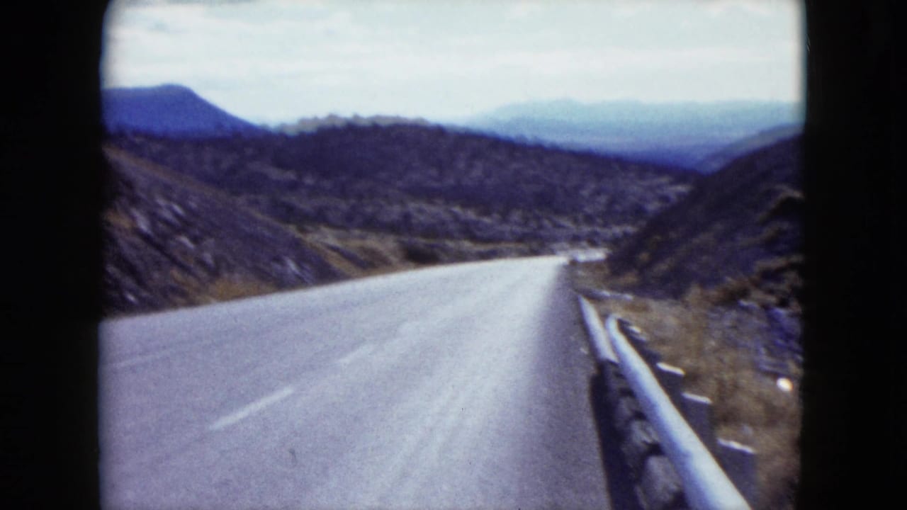 1959 Nevada home-movie frame looking down a paved road with a guardrail, dry hillsides, sparse shrubs, and distant mountains.
