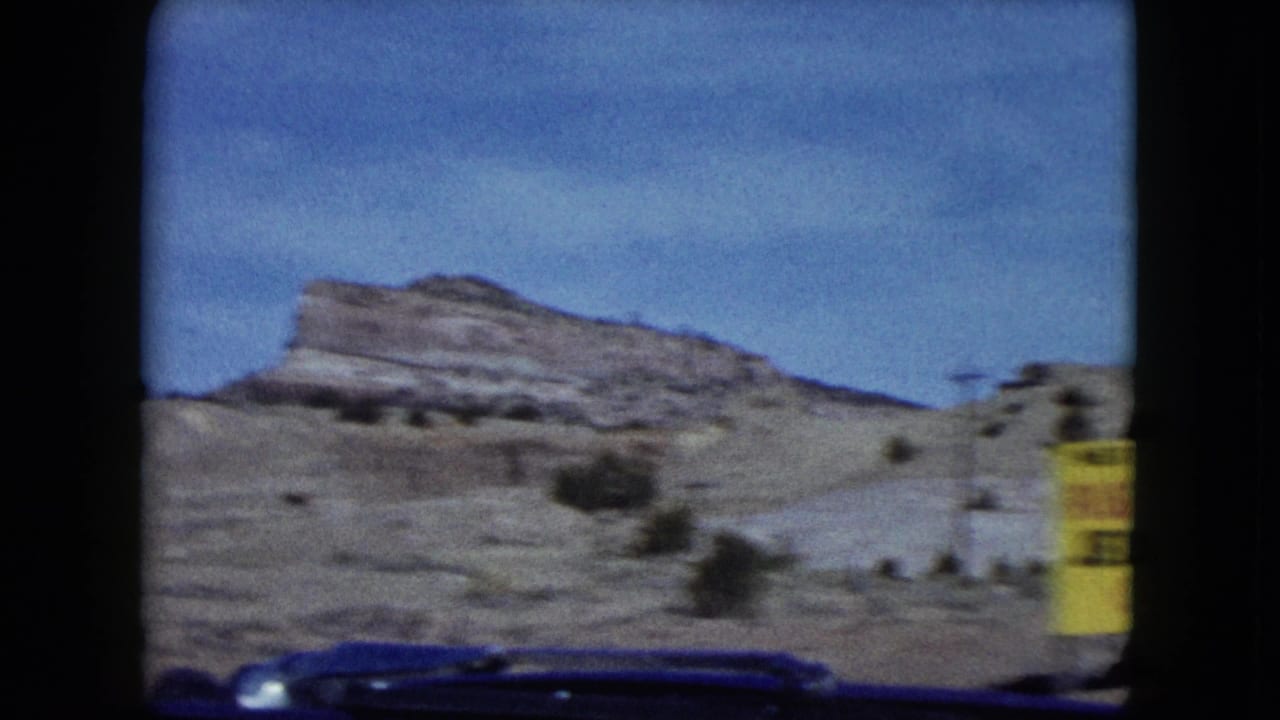 1959 Nevada 8mm road-trip frame showing rocky desert hills, scrub, a car hood, and a yellow roadside sign seen through a windshield.