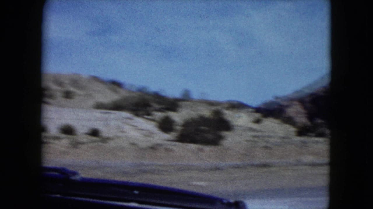 Roadside desert landscape with low rocky hills seen from inside a car.