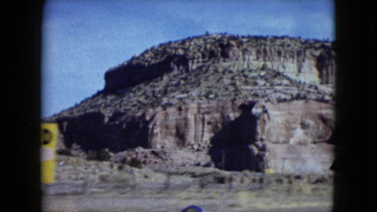 1959 Nevada 8mm frame showing a large rocky mesa, open desert ground, and the edge of a yellow roadside snack sign.