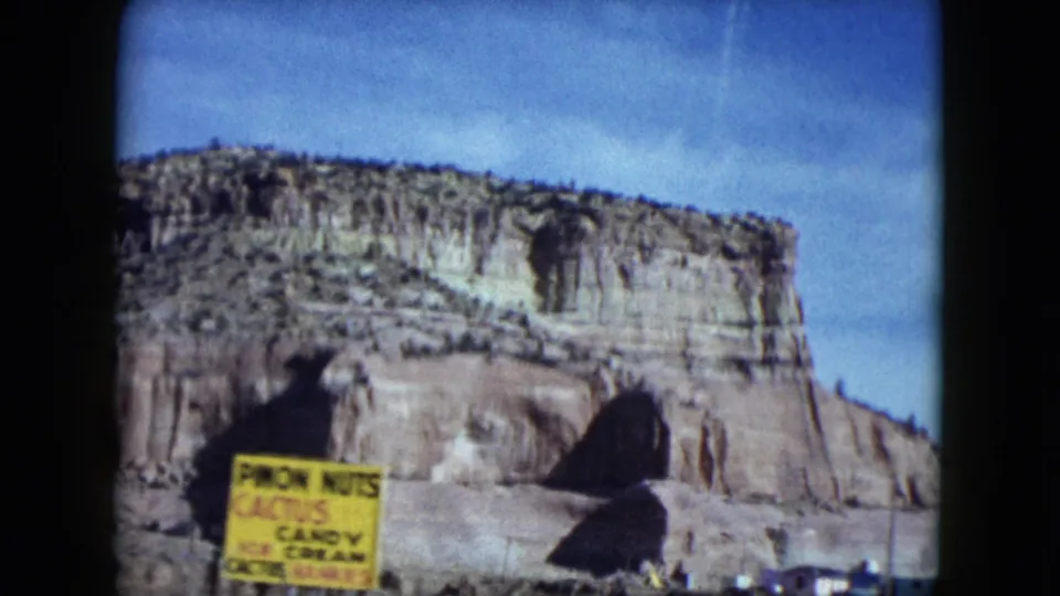 1959 Nevada road-trip frame with a rocky mesa behind a yellow sign reading pecan nuts, cactus candy, and ice cream.