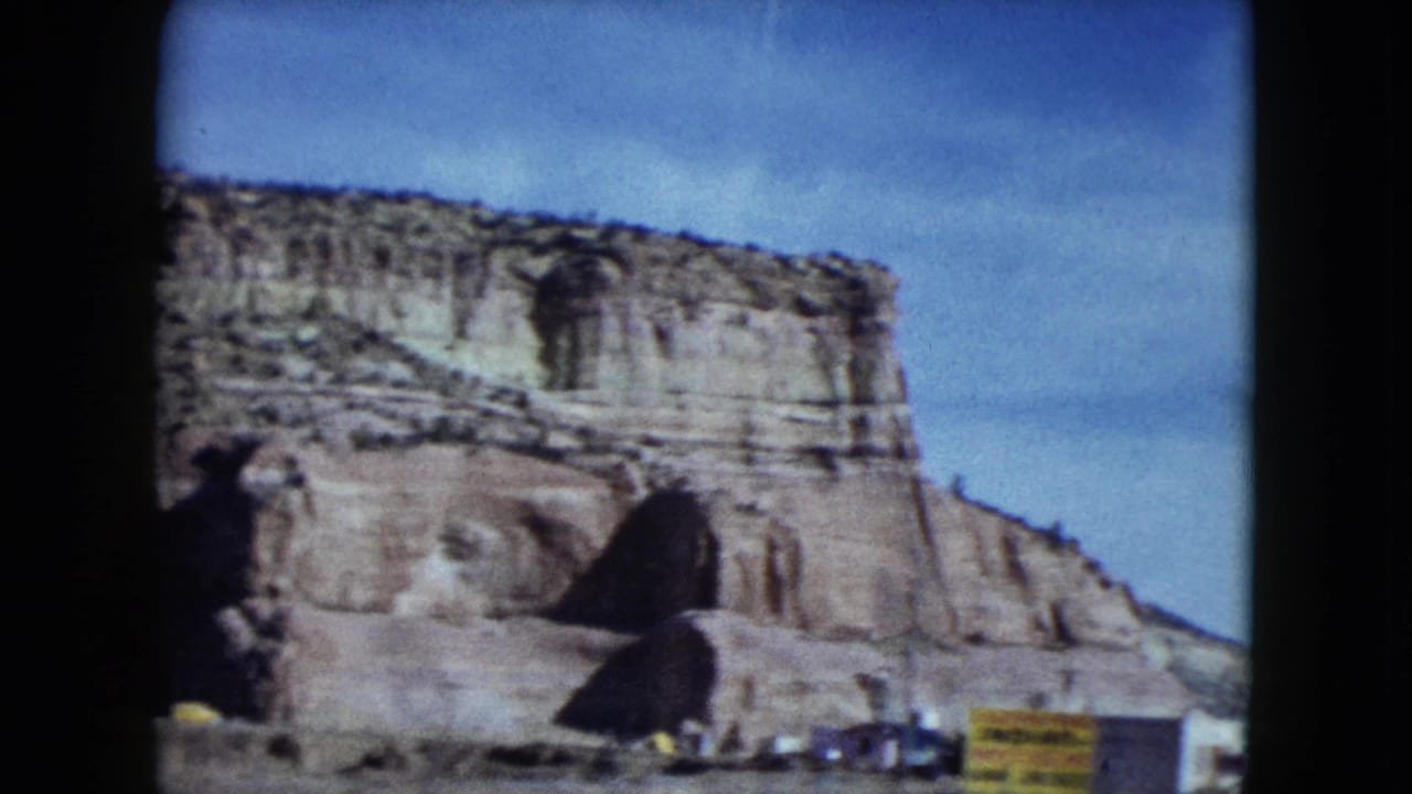 1959 Nevada road footage shows a sandstone cliff above small roadside signs and parked vehicles viewed from a moving car.