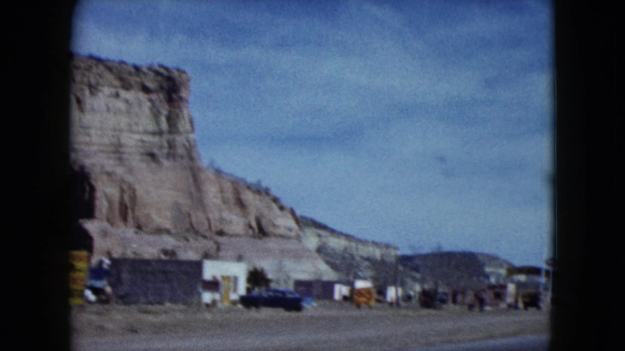 Roadside buildings and parked cars below a desert cliff.