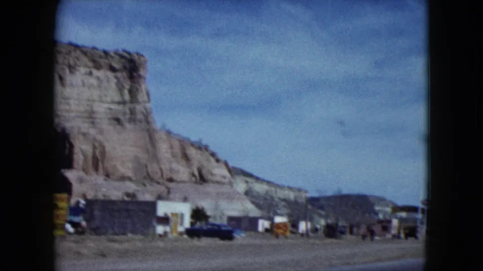 A Nevada roadside stop with low buildings, parked cars, signs, and a large sandstone cliff appears beside the road.
