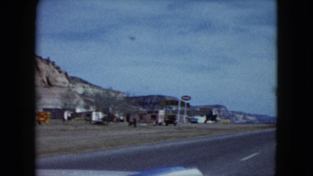 The moving-car view passes roadside businesses, parked vehicles, signs, and small distant people along a Nevada highway.