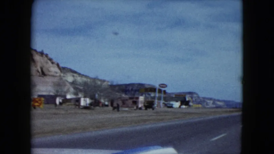 The moving-car view passes roadside businesses, parked vehicles, signs, and small distant people along a Nevada highway.