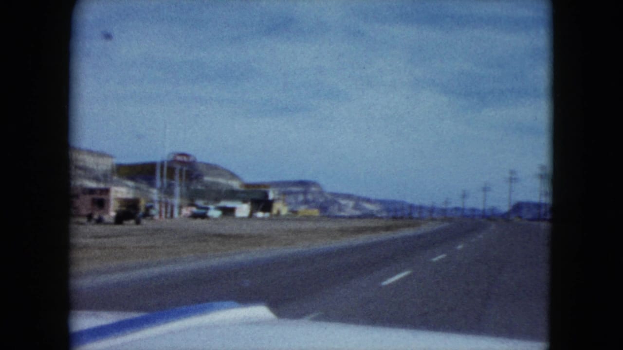 A forward-looking view from the car shows a paved Nevada highway with roadside buildings, signs, utility poles, and low desert hills.