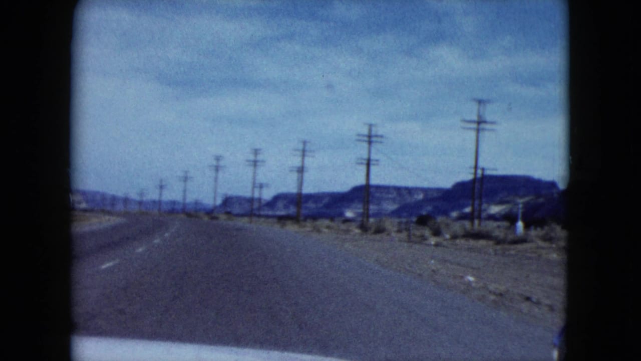 1959 Nevada highway footage looking ahead from a car toward low mesas, roadside poles and sparse desert shoulders.