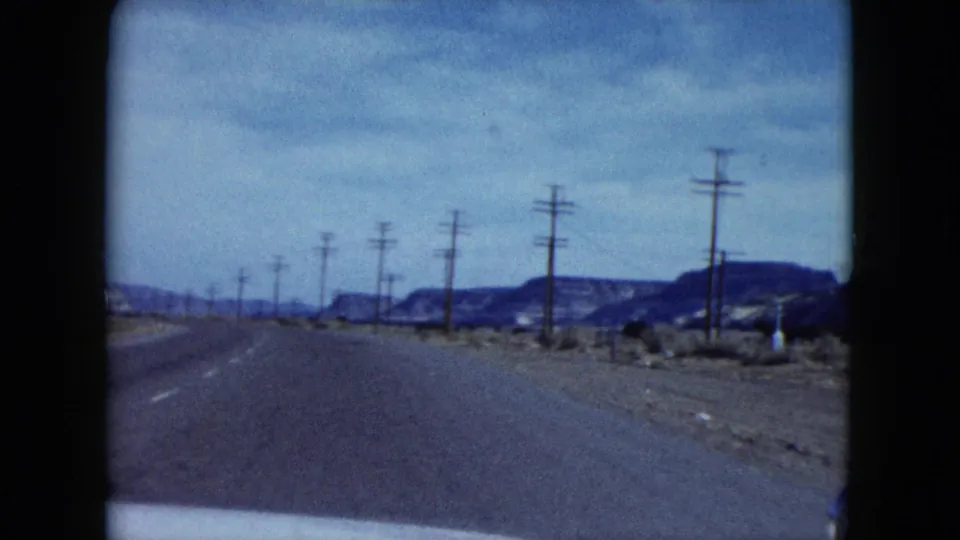 1959 Nevada highway footage looking ahead from a car toward low mesas, roadside poles and sparse desert shoulders.