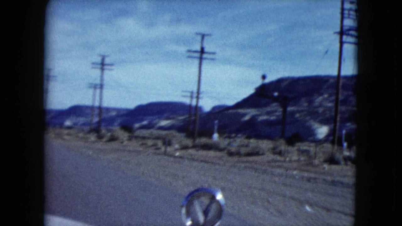 Moving-car view in Nevada, 1959, with roadside poles, scrubby desert ground, rocky hills and a small car-hood ornament visible.