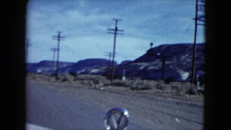 Moving-car view in Nevada, 1959, with roadside poles, scrubby desert ground, rocky hills and a small car-hood ornament visible.