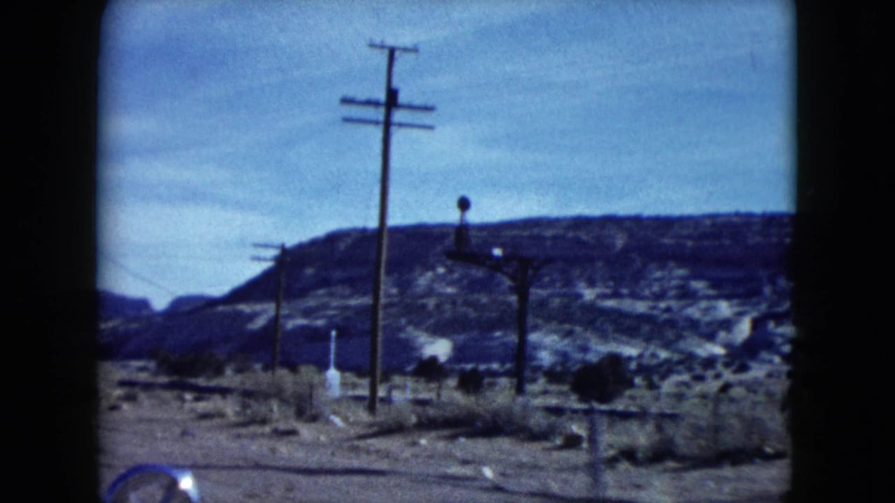 Nevada desert roadside in 1959 with a utility pole, small signpost, scrub ground and layered rocky mesas beyond.