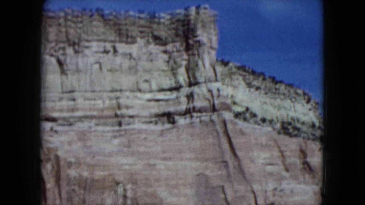 1959 Nevada travel footage frame showing a pale layered canyon wall, desert slope, and clear blue sky.