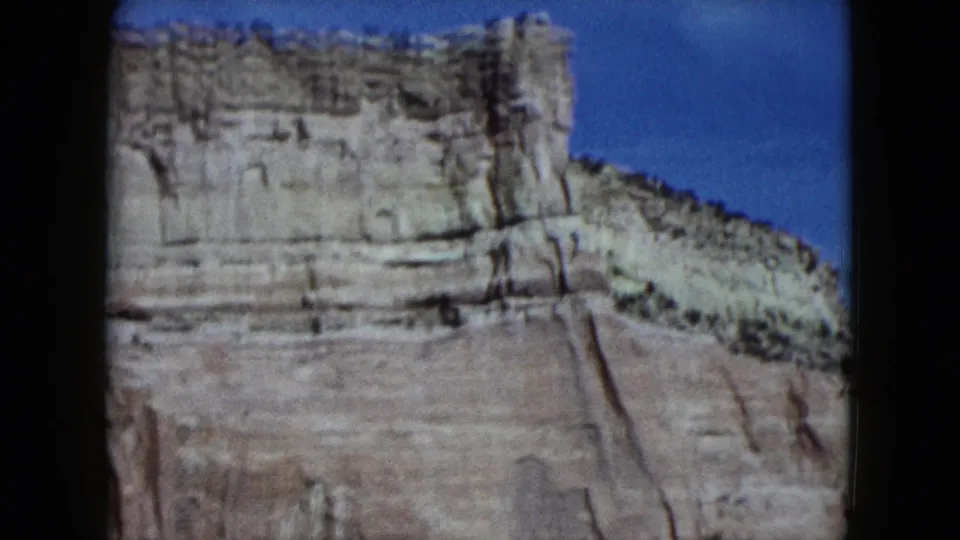 1959 Nevada travel footage frame showing a pale layered canyon wall, desert slope, and clear blue sky.