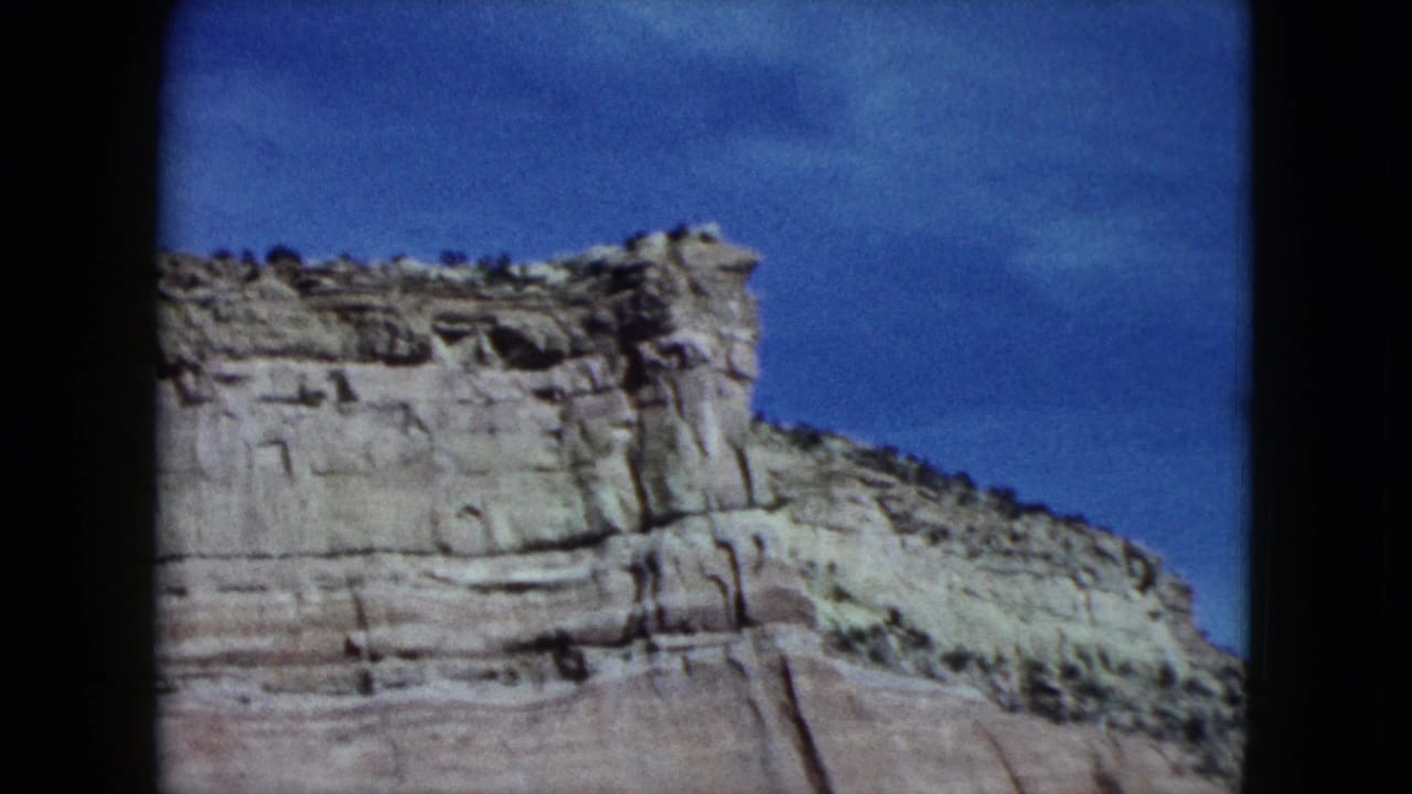 1959 Nevada 8mm frame showing a pale layered cliff face and sparse scrub beneath a blue sky from a moving-car viewpoint.