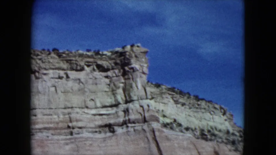 1959 Nevada 8mm frame showing a pale layered cliff face and sparse scrub beneath a blue sky from a moving-car viewpoint.