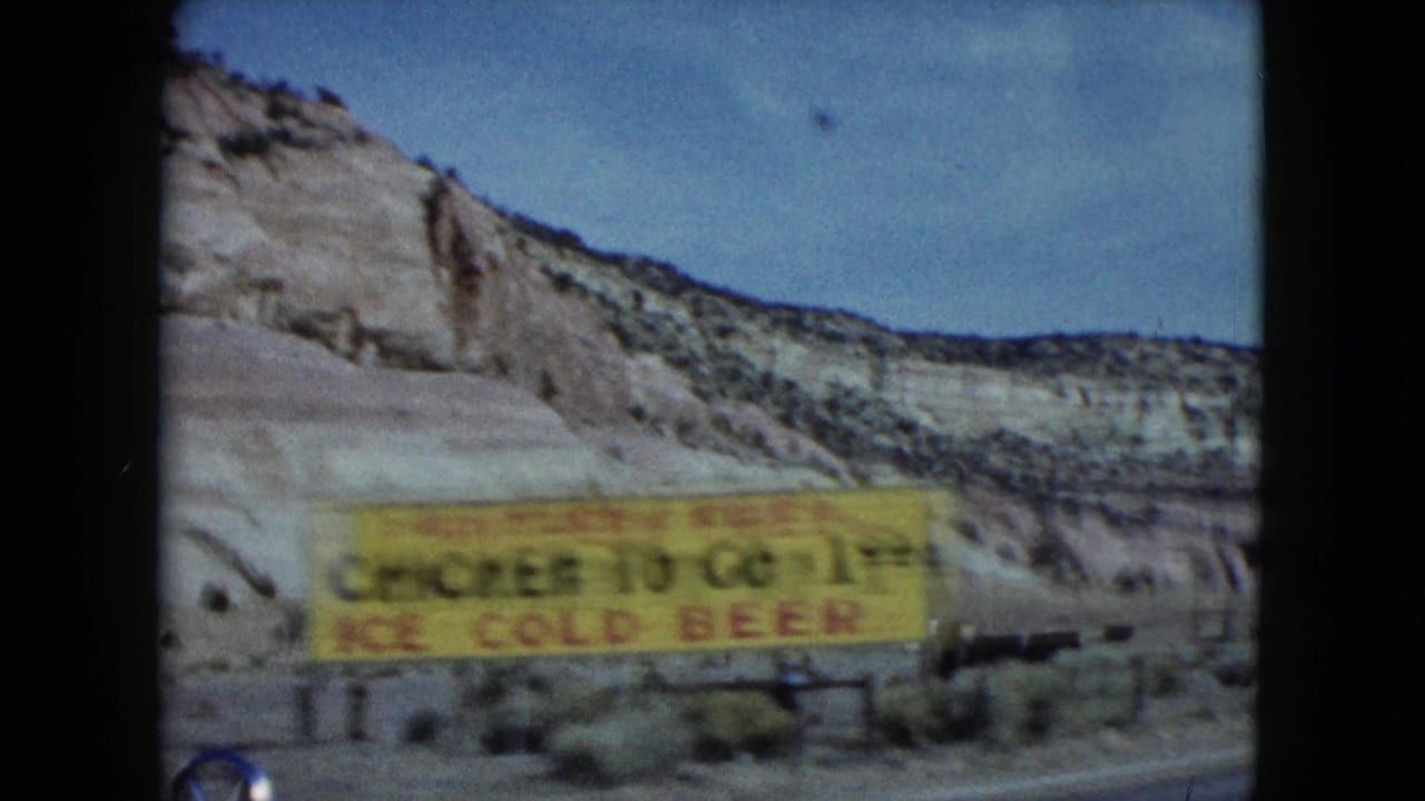 1959 Nevada 8mm frame showing a yellow roadside food sign passing in front of layered desert cliffs and scrub.