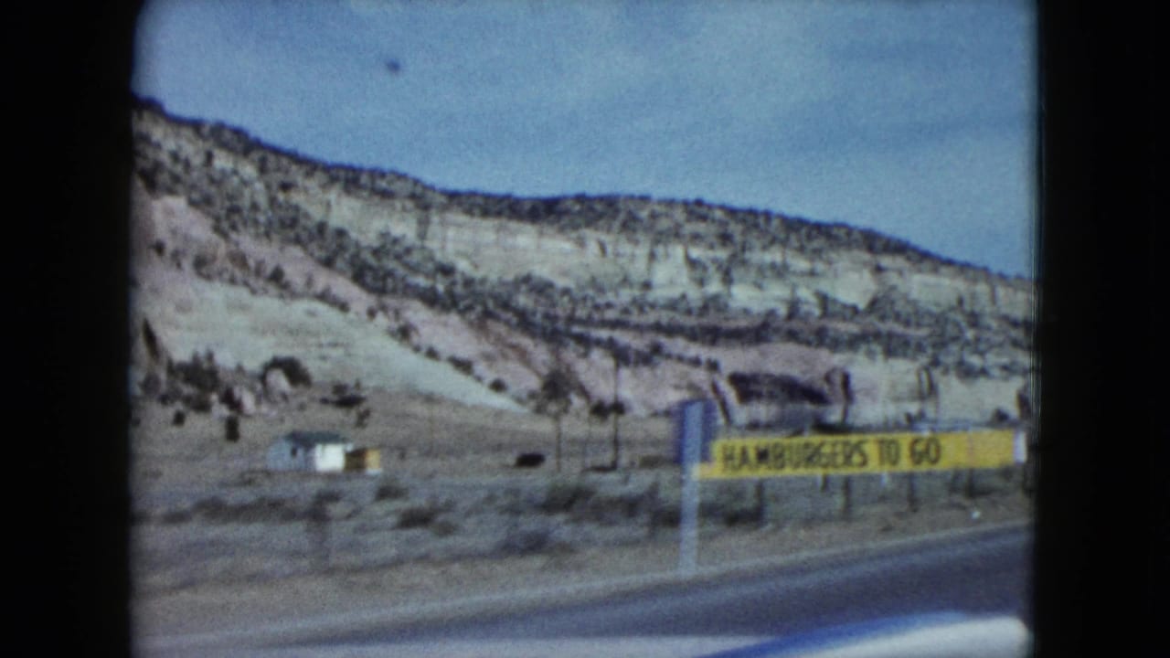 1959 Nevada frame showing a yellow sign reading hamburgers to go beside a road, with cliffs and utility poles in the background.