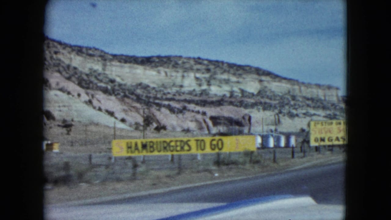 1959 Nevada road-trip frame with a yellow hamburgers-to-go sign, highway edge, roadside posts, and arid rock hills beyond.
