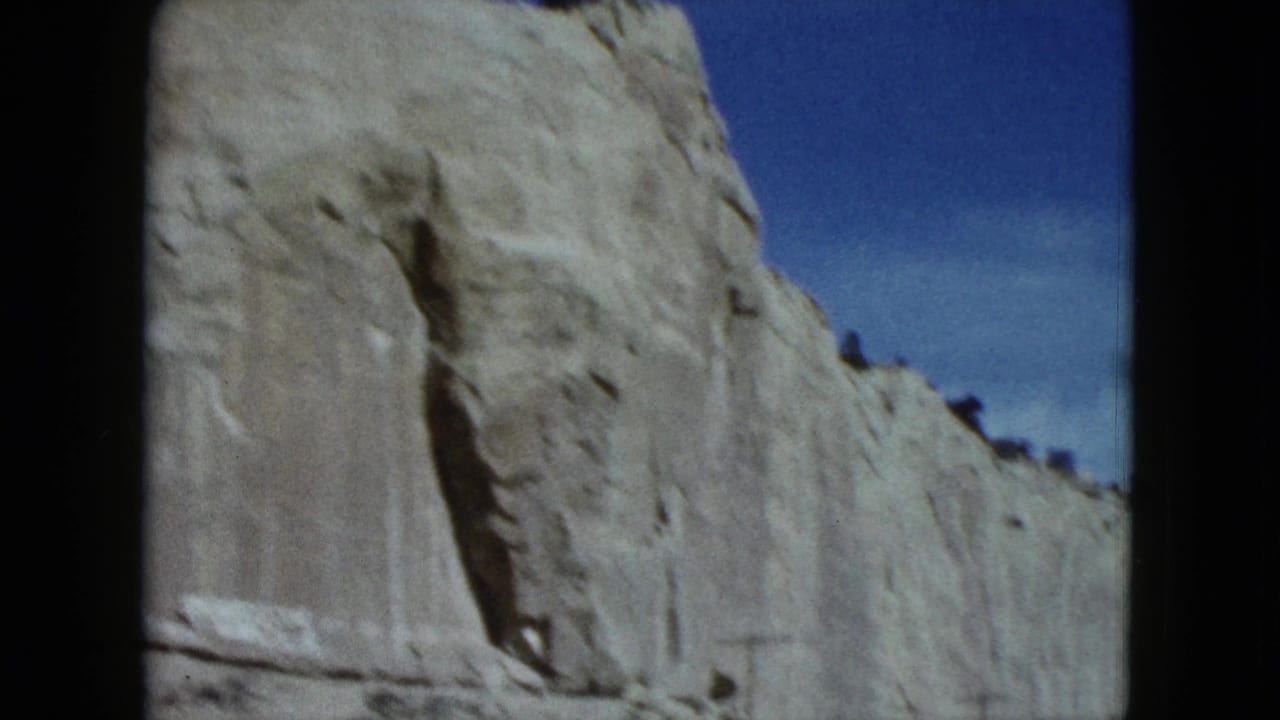 1959 Nevada road-trip frame showing a tall pale rock face, a vertical dark cleft, sparse ridge vegetation, and blue sky.