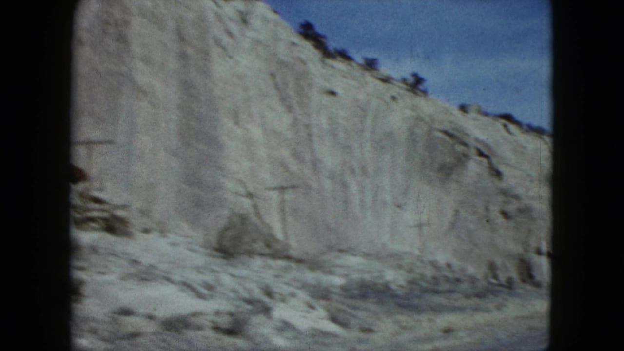 Final 1959 Nevada driving frame with an eroded pale cliff, scattered roadside rocks, ridge brush, and clear blue sky.