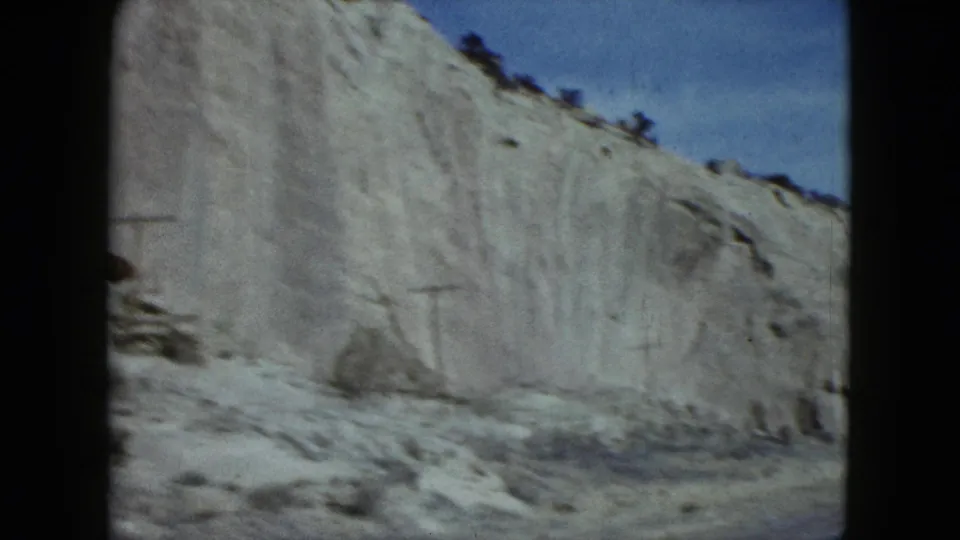 Final 1959 Nevada driving frame with an eroded pale cliff, scattered roadside rocks, ridge brush, and clear blue sky.