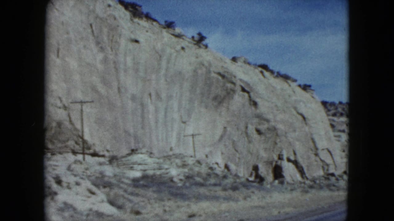 1959 Nevada 8mm frame showing a pale rock cliff, utility poles, dry roadside brush, and blue sky from a moving road trip.