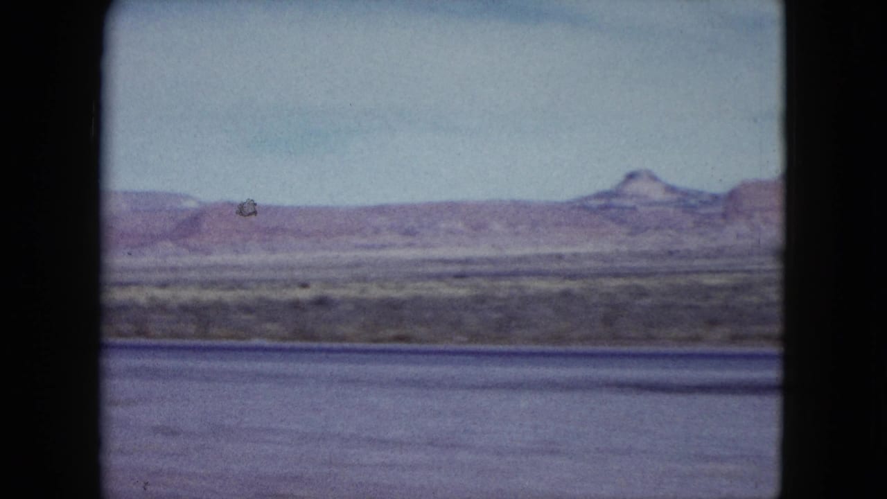 1959 Nevada landscape frame showing distant mesas, flat desert terrain, pale sky, and motion blur from the moving vehicle.