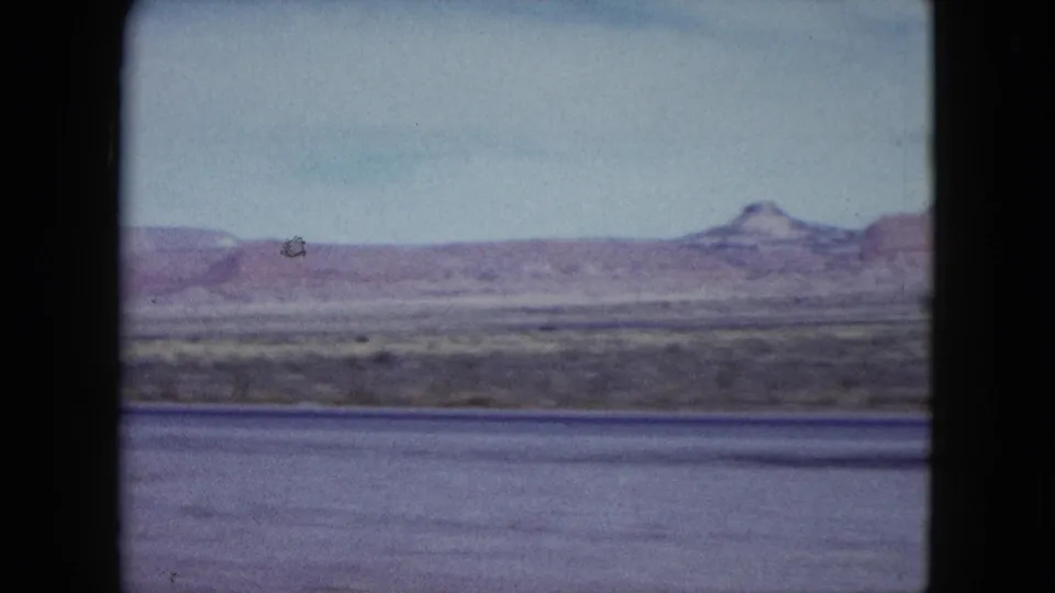 1959 Nevada landscape frame showing distant mesas, flat desert terrain, pale sky, and motion blur from the moving vehicle.