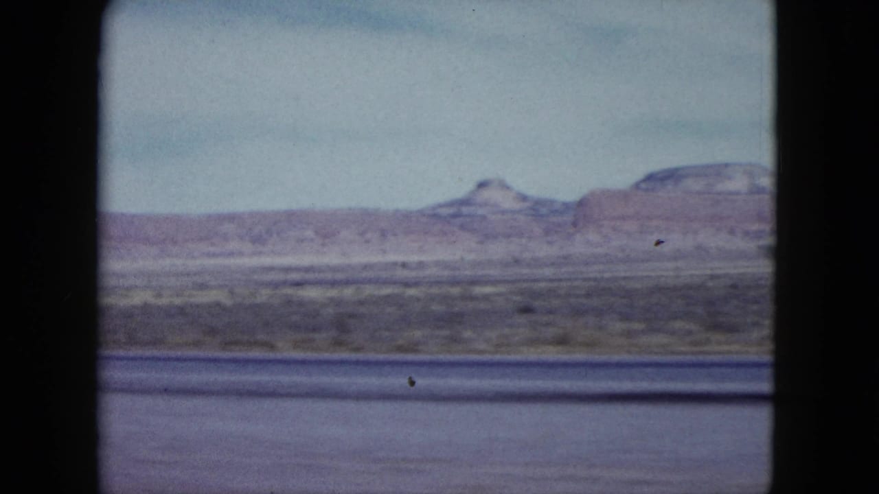 1959 Nevada desert plain with distant mesas and hazy sky, filmed from a moving vehicle on 8mm.
