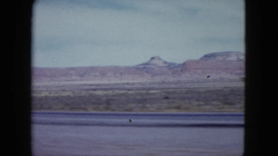 1959 Nevada desert plain with distant mesas and hazy sky, filmed from a moving vehicle on 8mm.