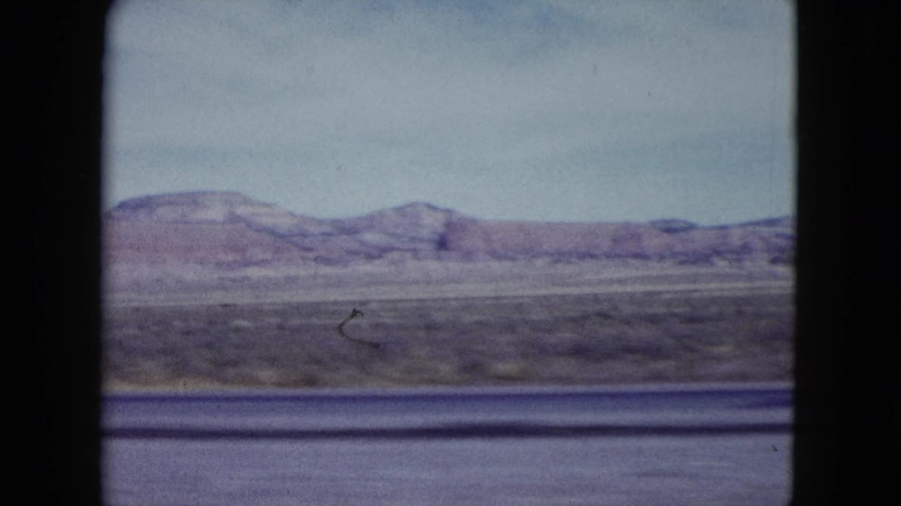 1959 Nevada roadside desert view with a low mesa ridge, scrub plain, and soft motion blur from travel.
