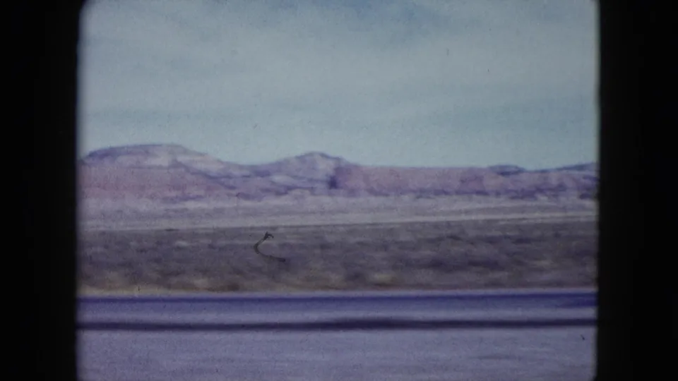 1959 Nevada roadside desert view with a low mesa ridge, scrub plain, and soft motion blur from travel.
