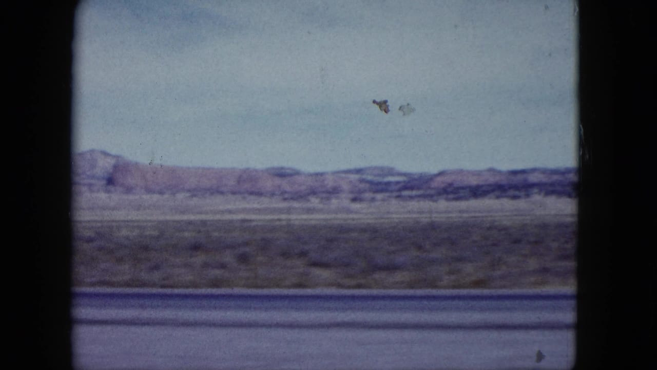 Nevada desert plain from a 1959 moving vehicle shot, with distant ridges and aged 8mm film texture.