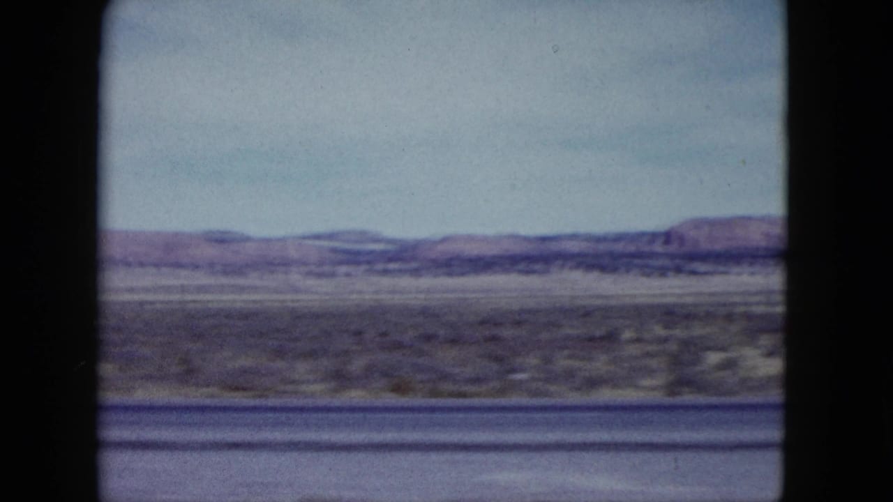 1959 Nevada home-movie frame showing arid roadside terrain, distant low mountains, pale sky, and dark car-window framing.
