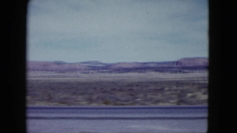 1959 Nevada home-movie frame showing arid roadside terrain, distant low mountains, pale sky, and dark car-window framing.