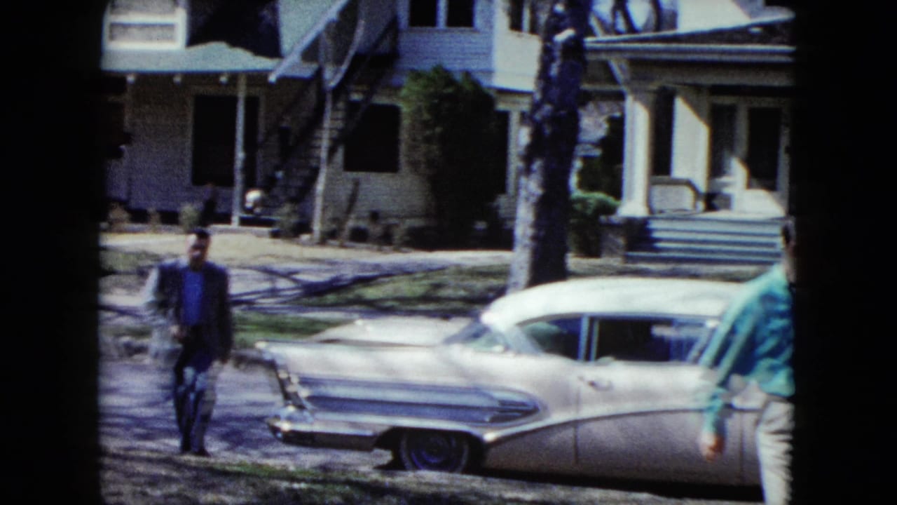 In this Nevada 1959 8mm frame, a man in a dark suit walks across a sunlit suburban lawn near a white 1950s sedan, with a two-story house and exterior stairs behind him.