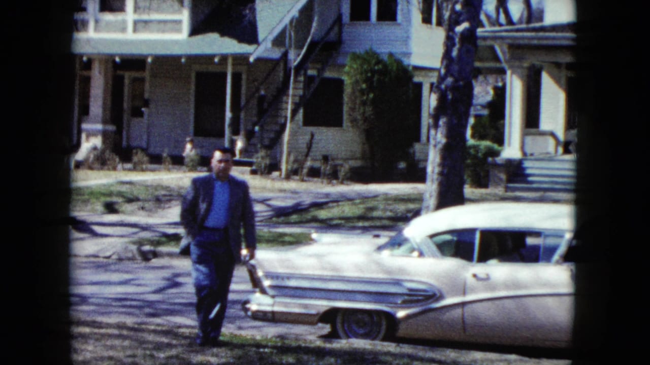 Man in a suit walks closer to a white sedan while another adult stands at the right edge.