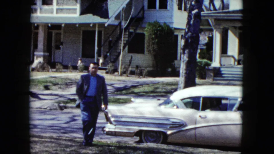 In this supported Nevada 1959 frame, the suited man walks toward the camera near the rear of a white finned sedan, with a sunlit house, lawn, tree, and a partially visible adult in green at the edge.