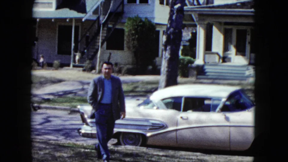 The Nevada 1959 home-movie frame shows the suited man nearer the foreground beside the white 1950s sedan, with the car hood open and a residential porch visible behind him.