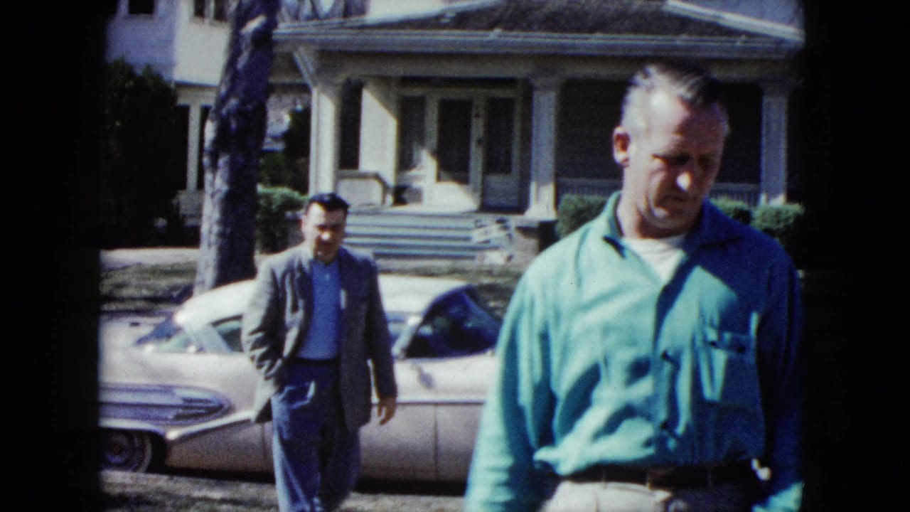 Two men walk near a pale car in front of a house.