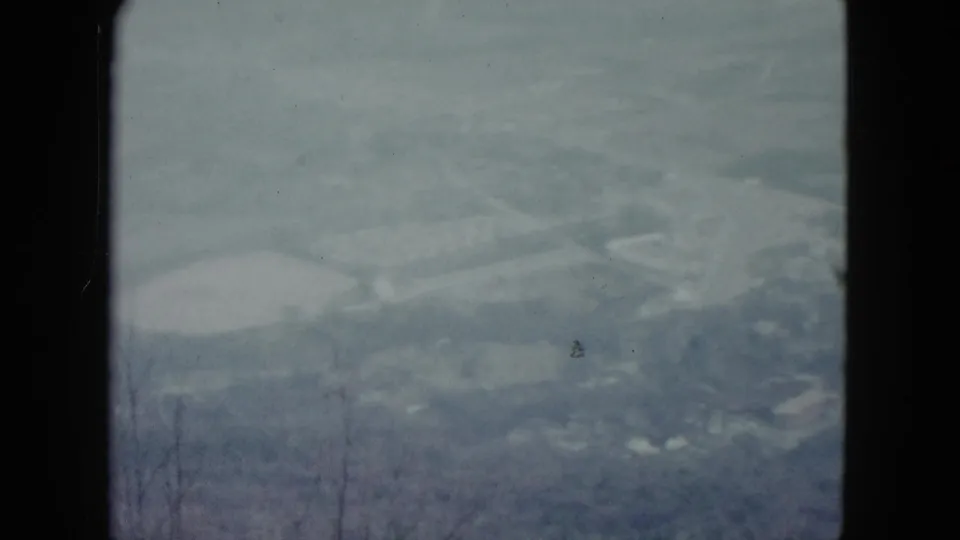 1959 Nevada 8mm still showing a hillside overlook toward a hazy valley, pale water feature, faint roads, and bare branch silhouettes.