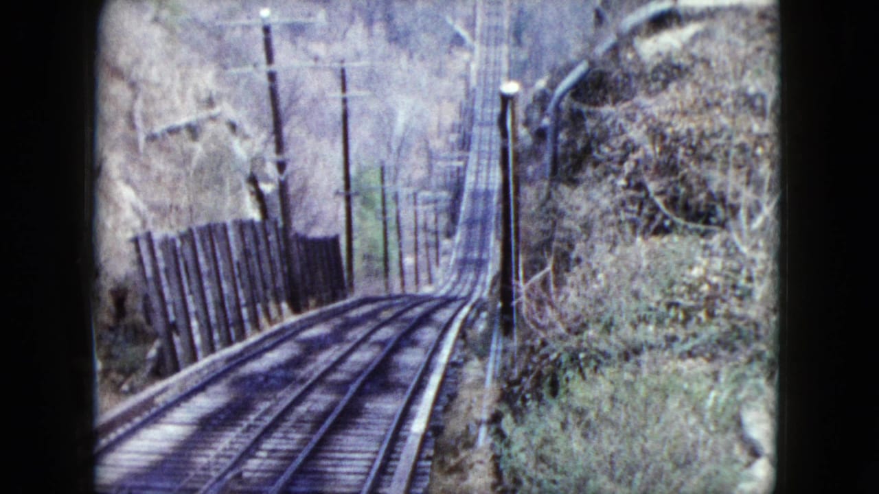 Curving train tracks descend past utility poles and brush on a wooded hillside.