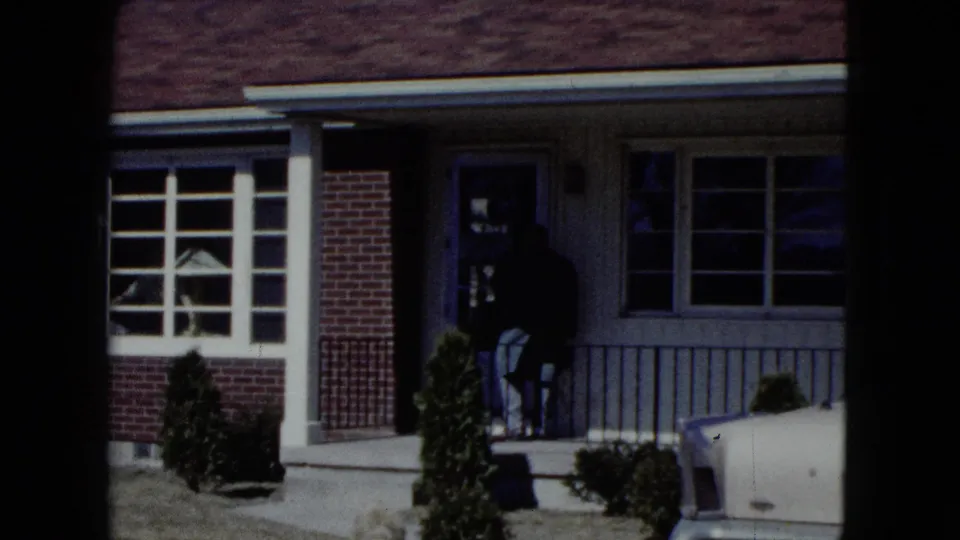 Older man in a dark suit appears at the front door of a Nevada suburban home in 1959 home-movie footage.
