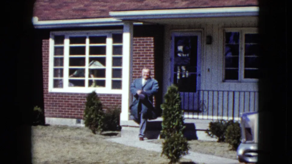 A smiling older man in a suit walks down the sidewalk from a Nevada suburban home, filmed on 8mm in 1959.