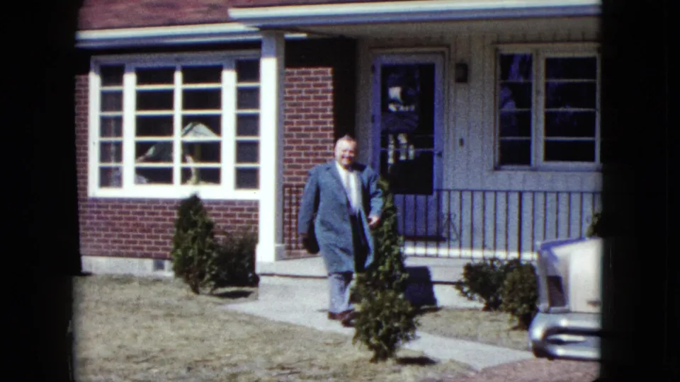 The suited man continues along the front walk outside the Nevada house, with shrubs and part of a car at the frame edge.
