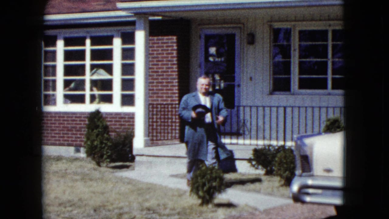 Man in a blue overcoat steps from a suburban house holding a hat.