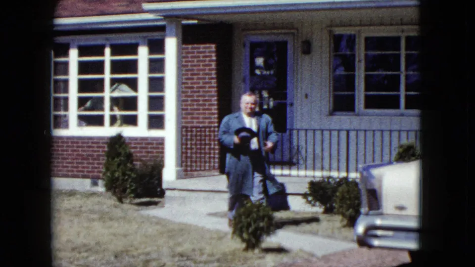 In Nevada 1959 home-movie footage, a well-dressed man walks from a brick-and-siding house while holding a brimmed hat.