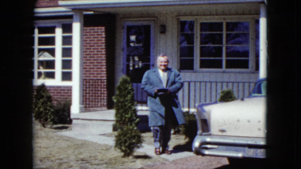 The man smiles toward the camera beside a pale 1950s car, with the Nevada suburban house and shrubs behind him.