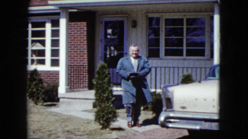 The man smiles toward the camera beside a pale 1950s car, with the Nevada suburban house and shrubs behind him.